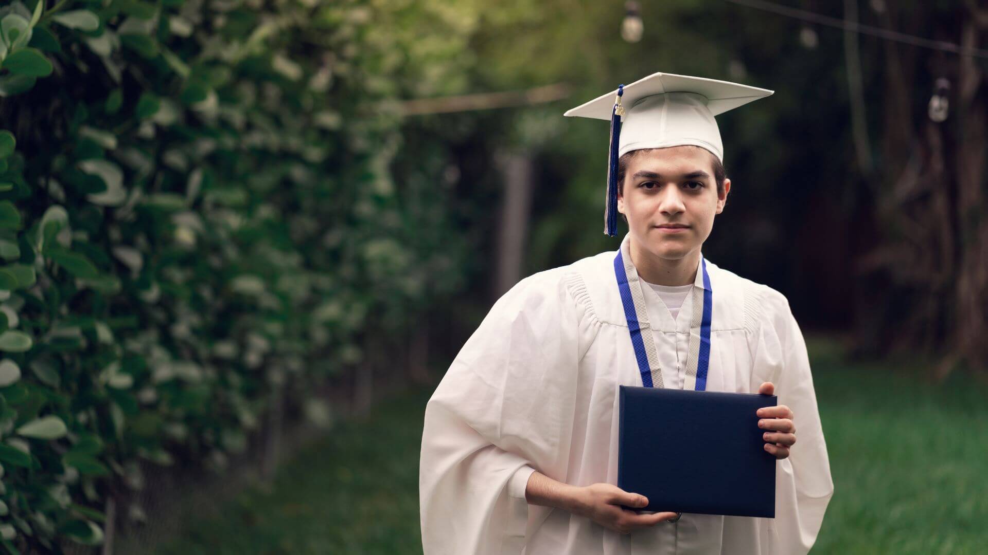 high school graduate in a cap and gown holding his high school diploma